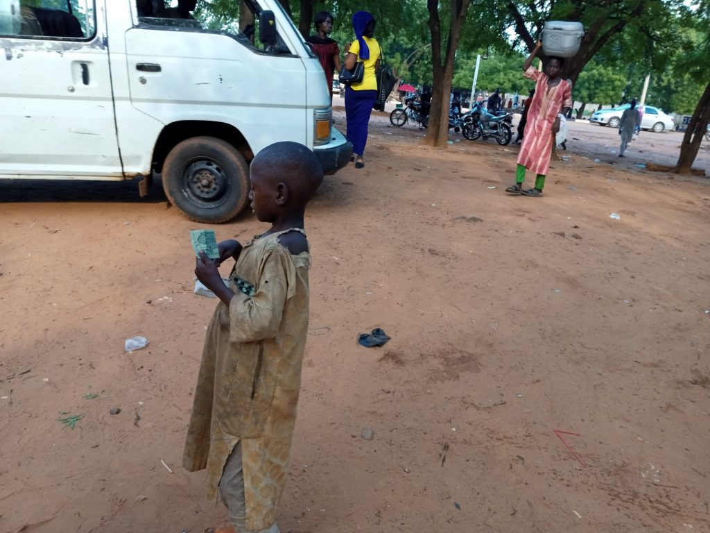 Almajiri child holding money on a street in Nigeria as other people move around nearby.