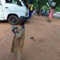 Almajiri child holding money on a street in Nigeria as other people move around nearby.