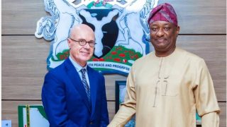 Nigerian Minister of Aviation Festus Keyamo wearing traditional attire shaking hands with a foreign official in front of the Nigerian Coat of Arms.