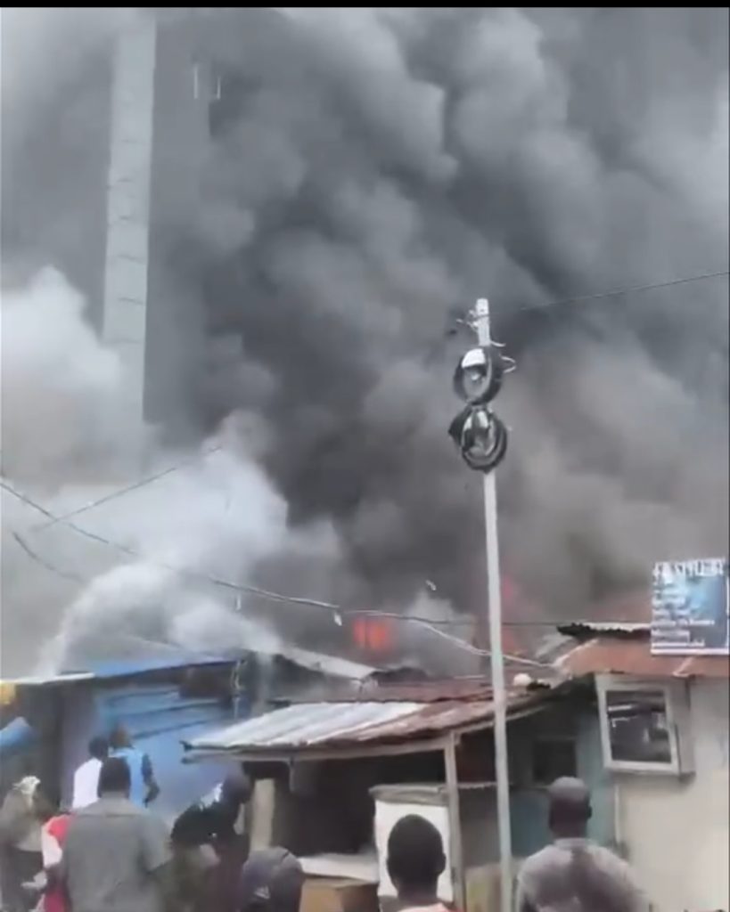 A dense column of dark smoke rises over low-rise structures along Abudu Street in Lagos, with bright flames glowing behind rooflines as local residents look on.